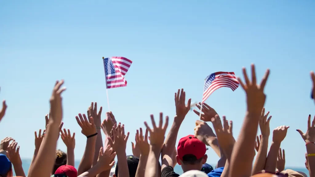 Crowd raising hands with American flags waving under a clear blue sky, celebrating Flag Day.