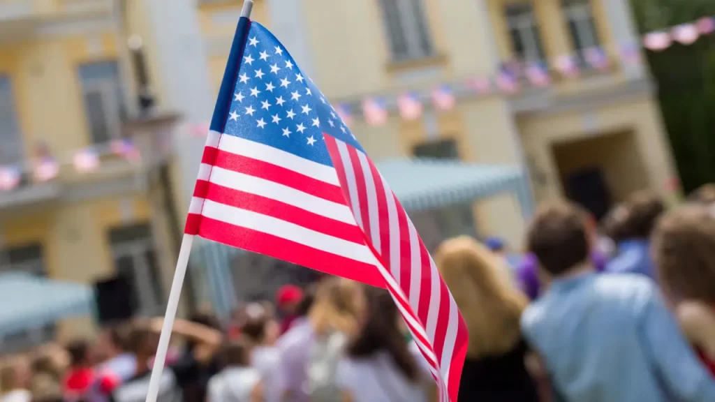 Close-up of an American flag on a stick with a crowd and decorated building in the background, celebrating Flag Day.